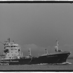 Schwarz‑weiß-Foto eines Frachtschiffs namens COMANCHE, seitliche Ansicht auf See; links unten Wasserzeichen des Internationalen Maritimen Museums Hamburg.