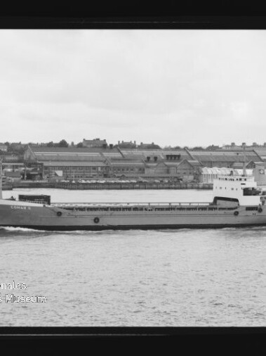 Flaches Frachtschiff fährt auf ruhigem Wasser vor einer Industriehafen-Kulisse mit Lagerhallen; links unten ist das Logo des Internationalen Maritimen Museums Hamburg zu sehen.