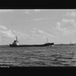 Schwarzweißaufnahme eines Frachtschiffs, das bei leicht bewegter See entlangfährt, mit Wellen im Vordergrund, wolkigem Himmel und einem kleinen Segelboot am rechten Rand; unten links das Logo des Internationalen Maritimen Museums Hamburg.
