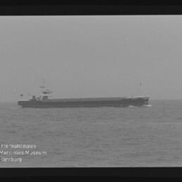 Schwarz-weiß-Foto eines einzelnen Frachtschiffs in Queransicht, das auf ruhiger See fährt, mit sichtbarem Filmrand und Stempel des Internationalen Maritimen Museums Hamburg.