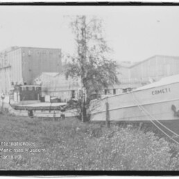 In Ufernähe vertäutes Schiff mit dem Namen COMET I vor Industriehallen, im Vordergrund ein Baum und grasbewachsenes Ufer.