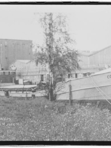 In Ufernähe vertäutes Schiff mit dem Namen COMET I vor Industriehallen, im Vordergrund ein Baum und grasbewachsenes Ufer.