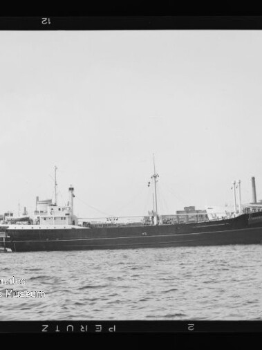 Schwarzweißaufnahme eines längsseits im Hafen liegenden Frachtschiffs in Seitenansicht mit Masten, Aufbauten und Schornstein vor Kaianlagen und Gebäuden, ruhige Wasserfläche im Vordergrund und sichtbare Filmrahmenkanten.