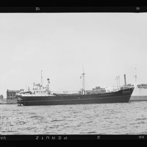 Schwarzweißaufnahme eines längsseits im Hafen liegenden Frachtschiffs in Seitenansicht mit Masten, Aufbauten und Schornstein vor Kaianlagen und Gebäuden, ruhige Wasserfläche im Vordergrund und sichtbare Filmrahmenkanten.
