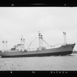 Schwarzweißfoto eines festlich beflaggten Frachtschiffs mit mehreren Masten und Ladekränen, das auf ruhigem Wasser vor flachem Küstenhintergrund fährt; am Bildrand sind Filmbandnummern und ein Wasserzeichen des Internationalen Maritimen Museums Hamburg sichtbar.