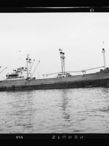 Schwarzweiß-Foto eines Frachtschiffs mit mehreren Ladebäumen und Aufbauten, das ruhig auf dem Wasser vor einem flachen Horizont liegt; unten links das Logo des Internationalen Maritimen Museums Hamburg.
