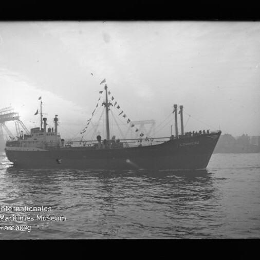 Schwarzweißaufnahme eines beflaggt geschmückten Handelsschiffs, das ruhig im Hafenwasser liegt, vor Kränen und Uferbebauung; unten links sichtbar das Wasserzeichen des Internationalen Maritimen Museums Hamburg.