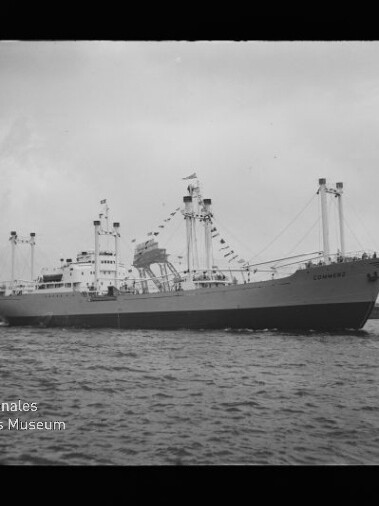 Großes, beflaggt geschmücktes Frachtschiff mit Ladekränen liegt seitlich im Hafen vor Industrieanlagen, Wasser im Vordergrund und Museumswasserzeichen unten links.