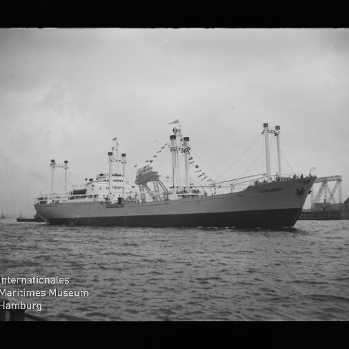 Großes, beflaggt geschmücktes Frachtschiff mit Ladekränen liegt seitlich im Hafen vor Industrieanlagen, Wasser im Vordergrund und Museumswasserzeichen unten links.