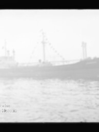 Verschwommene Silhouette eines Frachtschiffs mit Masten und einer Kette aus Signalflaggen auf ruhigem, nebligem Wasser, unten links ein rundes Museumswasserzeichen.