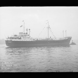 Schwarzweiß-Seitenansicht eines mit Signalfahnen geschmückten Frachtschiffs auf ruhigem Wasser, mit nebliger Küstenlinie im Hintergrund und kleinem Schleppboot am Heck.