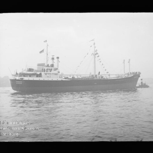 Schwarzweiß-Seitenansicht eines mit Signalfahnen geschmückten Frachtschiffs auf ruhigem Wasser, mit nebliger Küstenlinie im Hintergrund und kleinem Schleppboot am Heck.