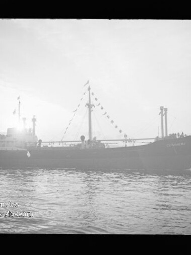 Schwarzweiß-Foto eines Frachtschiffs in Seitenansicht, silhouettenhaft vor hellem Himmel mit einer Girlande aus Signalflaggen am Mast, ruhigem Wasser im Vordergrund und Hafenkränen im Hintergrund; Wasserzeichen des Internationalen Maritimen Museums Hamburg unten links.