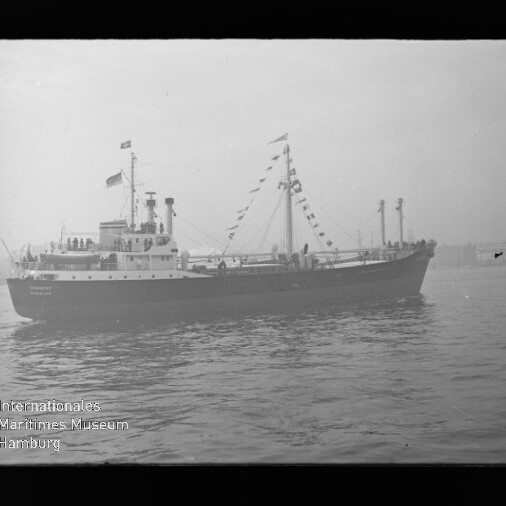 Schwarzweiß-Foto eines mit Signalfahnen geschmückten Frachtschiffs in Seitenansicht auf ruhigem Wasser, mit sichtbarer Brücke und Besatzung an Deck; Logowasserzeichen des Internationalen Maritimen Museums Hamburg unten links.