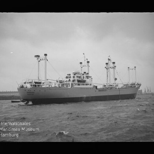 Schwarzweißaufnahme eines großen Frachtschiffs mit mehreren Ladebäumen und Aufbauten, das durch bewegtes Wasser vor einem Hafen mit Kranen fährt; unten links sichtbar das Logo des Internationalen Maritimen Museums Hamburg.