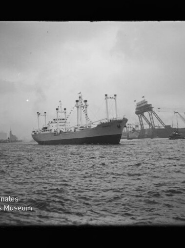 Mittelgroßes Frachtschiff mit mehreren Ladebäumen und Masten fährt auf welliger See vor einem Industriehafen mit hohen Krananlagen und Hafenbauten unter bedecktem Himmel.