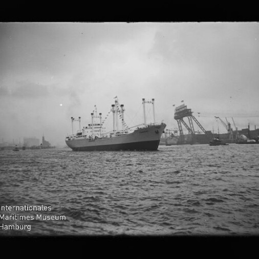 Mittelgroßes Frachtschiff mit mehreren Ladebäumen und Masten fährt auf welliger See vor einem Industriehafen mit hohen Krananlagen und Hafenbauten unter bedecktem Himmel.