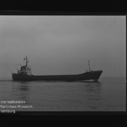 Schwarzweiß-Foto eines Frachtschiffs in Seitenansicht auf ruhiger See unter bedecktem Himmel; am unteren linken Bildrand sichtbar das Wasserzeichen „Internationales Maritimes Museum Hamburg“ sowie Filmränder mit Ziffern.
