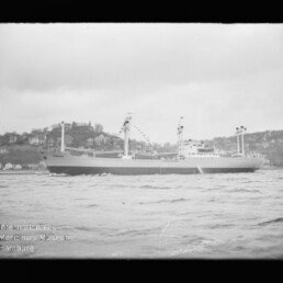 Schwarzweiß-Foto eines großen Frachtschiffs in Seitenansicht, geschmückt mit Signalfahnen und auf leichten Wellen fahrend vor einer hügeligen, mit Häusern bebauten Küste; unten links das Logo des Internationalen Maritimen Museums Hamburg.