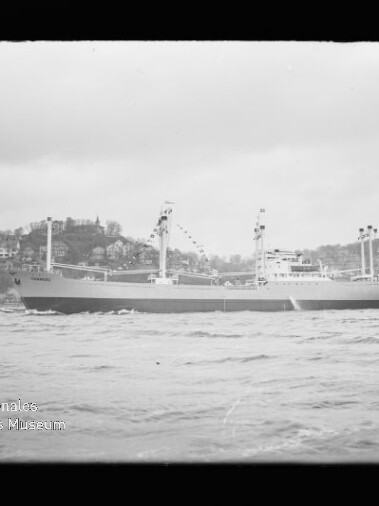 Schwarzweiß-Foto eines großen Frachtschiffs in Seitenansicht, geschmückt mit Signalfahnen und auf leichten Wellen fahrend vor einer hügeligen, mit Häusern bebauten Küste; unten links das Logo des Internationalen Maritimen Museums Hamburg.