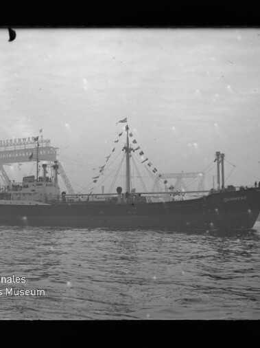 Schwarz-weiß-Foto eines beflagten Frachtschiffs in seitlicher Ansicht auf ruhiger See, mit Hafenbauten im Hintergrund und Wasserzeichen des Internationalen Maritimen Museums Hamburg.