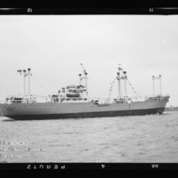 Schwarzweiß-Fotografie eines mittelgroßen Frachtschiffs mit mehreren Ladekränen und einer Girlande aus Signalflaggen, das auf ruhiger See vor einer flachen Küstenlinie fährt; Filmrand und Museumswasserzeichen sichtbar.