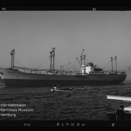 Schwarzweißaufnahme eines großen Frachtschiffs mit Ladekränen und Masten im Hafen, im Vordergrund ein kleines Boot auf dem Wasser und rechts die Silhouette einer Person an einer Pier; unten links das Logo des Internationalen Maritimen Museums Hamburg.