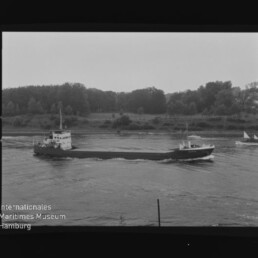 Schwarzweißfotografie eines langen Frachtschiffs, das auf einem Fluss vor einem baumbestandenen Ufer fährt; rechts im Bild ist ein kleines Segelboot zu sehen und unten links das Logo des Internationalen Maritimen Museums Hamburg.