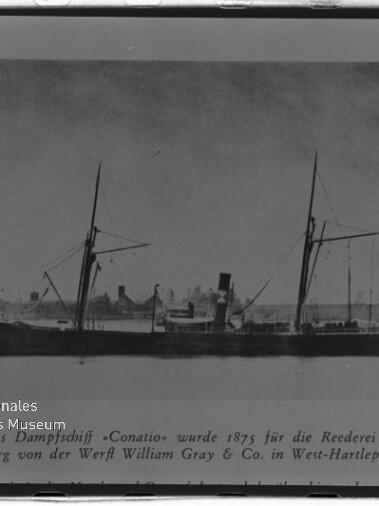Seitliche Schwarzweißaufnahme eines historischen Dampfschiffs mit zwei Masten und Schornstein auf ruhigem Wasser, im Hintergrund flaches Ufer mit Gebäuden und unten links ein Museumslogo und Schriftzug.