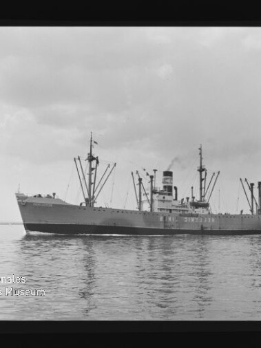 Schwarzweißaufnahme eines Frachtschiffs mit mehreren Ladebäumen und einem Schornstein, das ruhig über spiegelndes Wasser fährt; Filmrand mit KODAK-Markierung und Wasserzeichen des Internationalen Maritimen Museums Hamburg sichtbar.