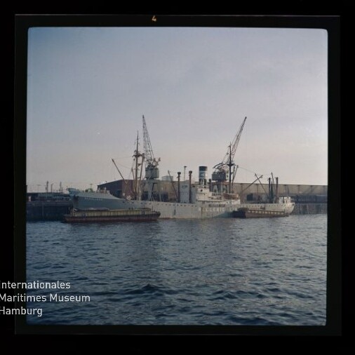 Großes graues Schiff liegt am Kai eines Industriehafens, flankiert von zwei Schleppern und mehreren Kranauslegern, ruhiges Hafenwasser im Vordergrund und ein Museum-Logo mit Schriftzug unten links