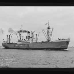 Schwarzweißfoto eines Frachtschiffs der Hellenic Lines mit mehreren Ladebäumen und rauchendem Schornstein, das auf ruhiger See fährt; unten links ist ein Wasserzeichen des Internationalen Maritimen Museums Hamburg zu sehen.