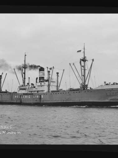 Schwarzweißfoto eines Frachtschiffs der Hellenic Lines mit mehreren Ladebäumen und rauchendem Schornstein, das auf ruhiger See fährt; unten links ist ein Wasserzeichen des Internationalen Maritimen Museums Hamburg zu sehen.