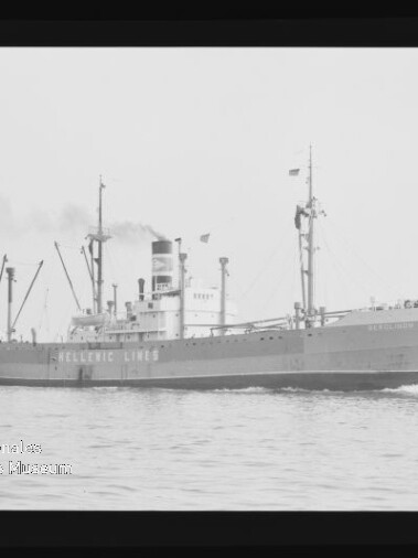 Schwarzweiß-Foto eines Frachtschiffs der Reederei Hellenic Lines mit hohem Schornstein, mehreren Ladekränen und Flaggen, das bei ruhiger See eine leichte Bugwelle zieht.