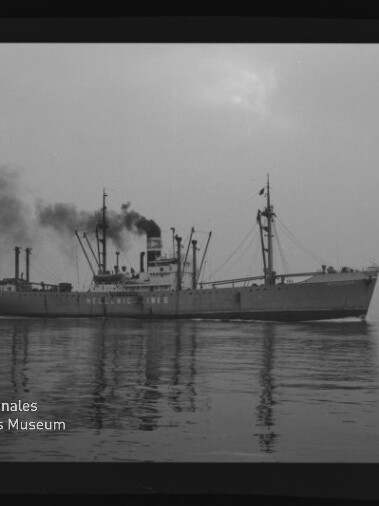 Schwarzweiß-Foto eines Frachtschiffs mit rauchendem Schornstein, Ladekränen und Spiegelung auf ruhiger See; unten links das Siegel des Internationalen Maritimen Museums Hamburg.