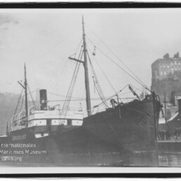 Schwarzweiß-Aufnahme eines im Hafen angelegten Dampfschiffs mit Schornstein, Masten und Ladegeschirr, rechts Kai mit Personen und Gebäuden auf einer Anhöhe; unten links sichtbar das Logo des Internationalen Maritimen Museums Hamburg.