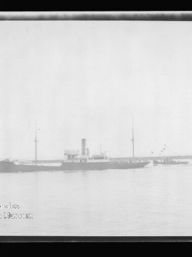 Schwarzweißaufnahme eines langgestreckten Frachtschiffs mit Schornstein und zwei Masten, begleitet von einem Schlepper, vor einer Hafenmole mit Leuchtturm auf ruhigem Wasser; Museumswasserzeichen unten links.
