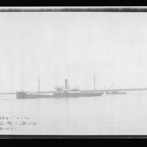 Schwarzweißaufnahme eines langgestreckten Frachtschiffs mit Schornstein und zwei Masten, begleitet von einem Schlepper, vor einer Hafenmole mit Leuchtturm auf ruhigem Wasser; Museumswasserzeichen unten links.