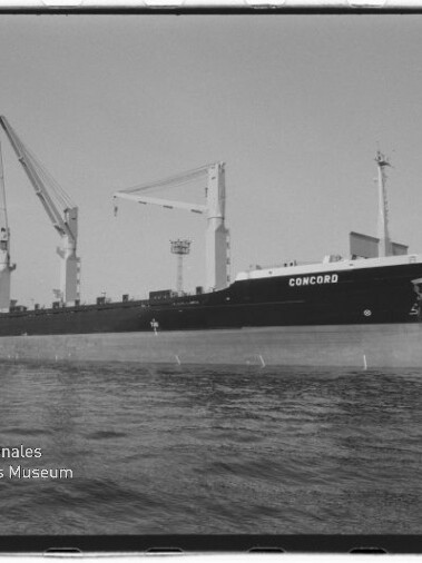 Schwarzweiß-Foto eines Frachtschiffs mit dem Namen CONCORD, längsseits im Hafen liegend mit mehreren Ladekränen an Deck, Hafenanlagen im Hintergrund und Wasser im Vordergrund, im unteren linken Eck das Logo des Internationalen Maritimen Museums Hamburg.