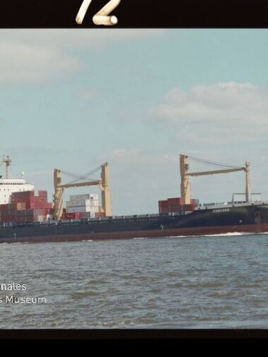 Containerfrachtschiff mit gestapelten Containern und zwei großen Ladekränen fährt auf ruhiger See unter hellem, leicht bewölktem Himmel; links unten Wasserzeichen des Internationalen Maritimen Museums Hamburg.