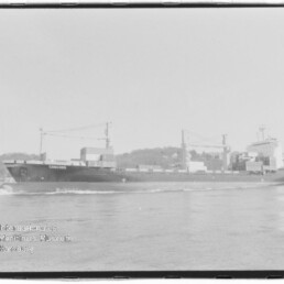 Schwarzweißaufnahme eines Containerschiffs mit dem Namen CONCORD, das auf ruhigem Wasser an einem bebaumten Ufer mit Hafenkränen und Stapelcontainern vorbeifährt.