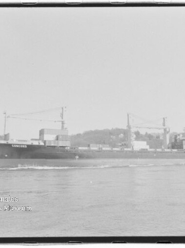 Schwarzweißaufnahme eines Containerschiffs mit dem Namen CONCORD, das auf ruhigem Wasser an einem bebaumten Ufer mit Hafenkränen und Stapelcontainern vorbeifährt.