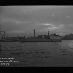 Schwarzweiß-Aufnahme eines Frachtschiffs im Hafen bei Dämmerung mit welligem Wasser im Vordergrund, Kränen und Hafenbauten im Hintergrund und dem Logo/Schriftzug des Internationalen Maritimen Museums Hamburg unten links.