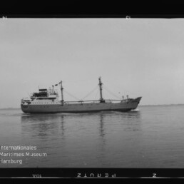 Schwarzweißaufnahme eines Frachtschiffs in Seitenansicht mit Ladebäumen und Aufbauten, das ruhig auf ruhiger See fährt; am unteren linken Rand das Logo des Internationalen Maritimen Museums Hamburg sowie Filmrand und -zahlen sichtbar.