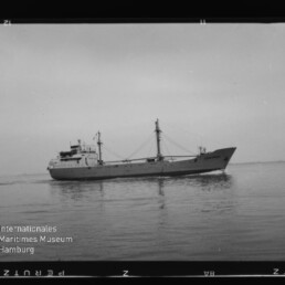 Schwarzweißfoto eines Frachtschiffs (Seitenansicht, Name „CONCORDIA“ am Bug) auf ruhiger See mit sichtbaren Masten, Ladegeschirr und leichtem Spiegelbild im Wasser; Filmrand und Museumswasserzeichen unten links erkennbar.