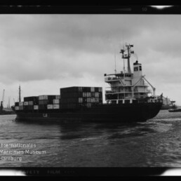 Schwarzweiß-Fotografie eines Containerschiffs im Hafen mit gestapelten Frachtcontainern und markantem Steuerhaus am Heck, ruhiger Wasseroberfläche und wolkenverhangenem Himmel; unten links Logo des Internationalen Maritimen Museums Hamburg.