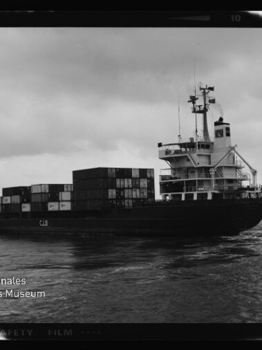 Schwarzweiß-Fotografie eines Containerschiffs im Hafen mit gestapelten Frachtcontainern und markantem Steuerhaus am Heck, ruhiger Wasseroberfläche und wolkenverhangenem Himmel; unten links Logo des Internationalen Maritimen Museums Hamburg.