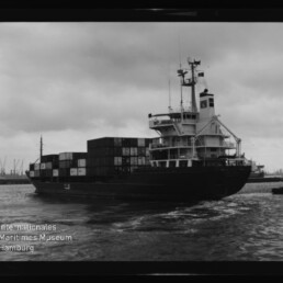 Ein Containerschiff mit gestapelten Ladecontainern und markantem Brückenaufbau fährt in einem Hafen bei bewölktem Himmel; unten links ist ein Wasserzeichen des Internationalen Maritimen Museums Hamburg zu sehen.
