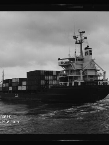 Ein Containerschiff mit gestapelten Ladecontainern und markantem Brückenaufbau fährt in einem Hafen bei bewölktem Himmel; unten links ist ein Wasserzeichen des Internationalen Maritimen Museums Hamburg zu sehen.