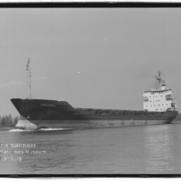 Schwarzweißaufnahme eines Frachtschiffs mit dem Namen CONCORDIA, seitliche Ansicht beim Fahren auf ruhigem Wasser mit langem Laderaum und Aufbauten achtern; Ufer mit Bäumen im Hintergrund und Museumswasserzeichen unten links.